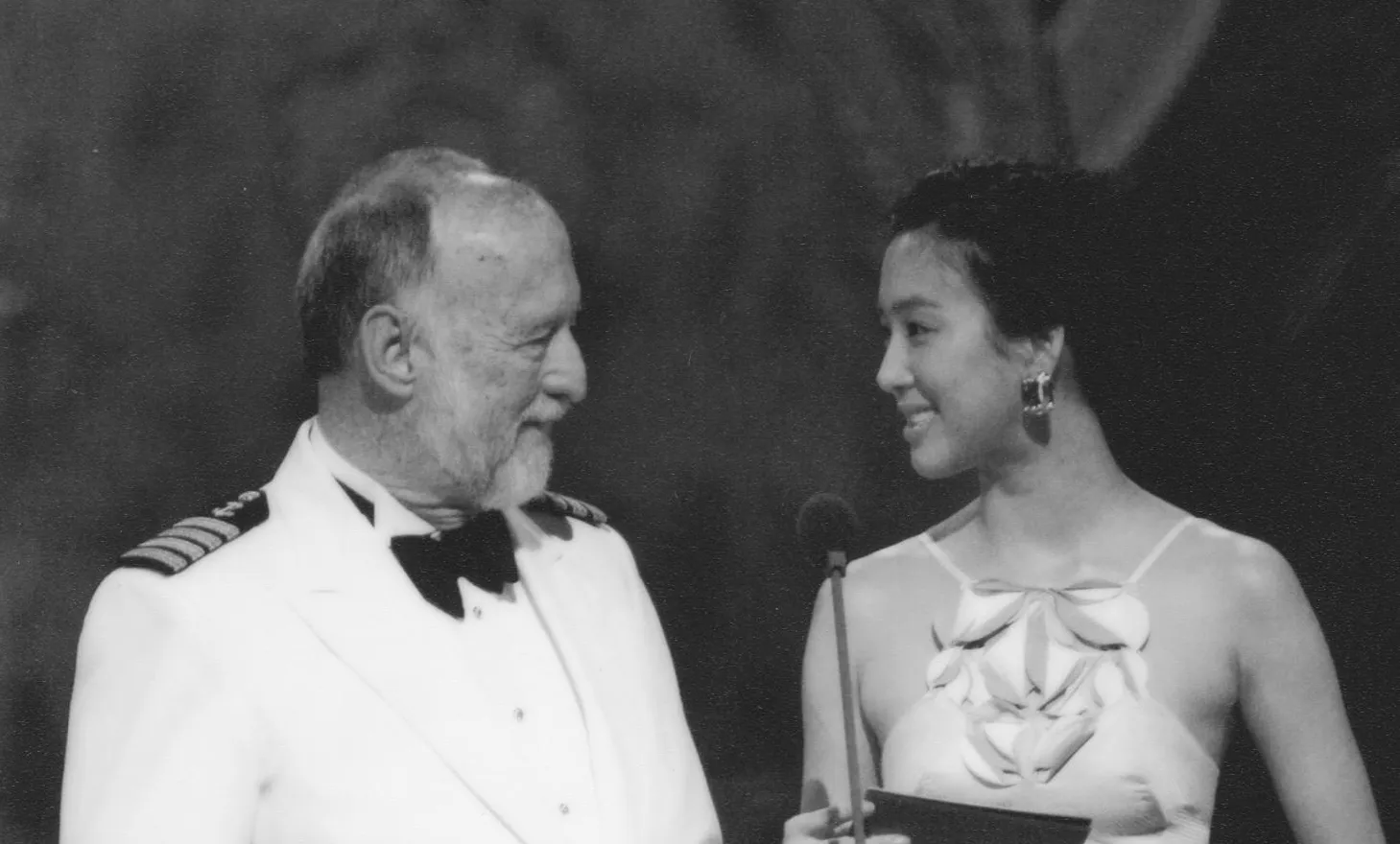 Black and white photo of an older man in a white uniform with a bow tie and a younger woman in an elegant dress smiling at each other on a stage with a microphone between them.
