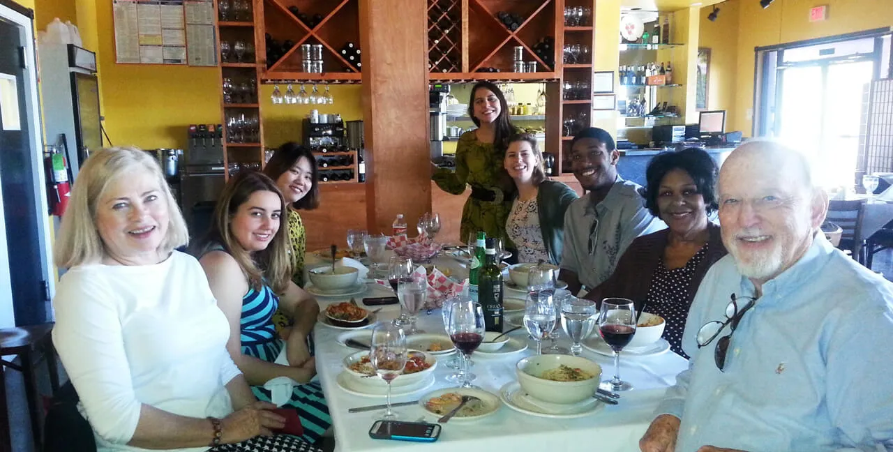 Eight people, a mix of men and women of diverse ages and ethnicities, smiling and seated around a white-tableclothed table with plates, bowls, wine glasses, and bottles in a cozy restaurant setting with wooden shelves and yellow walls.