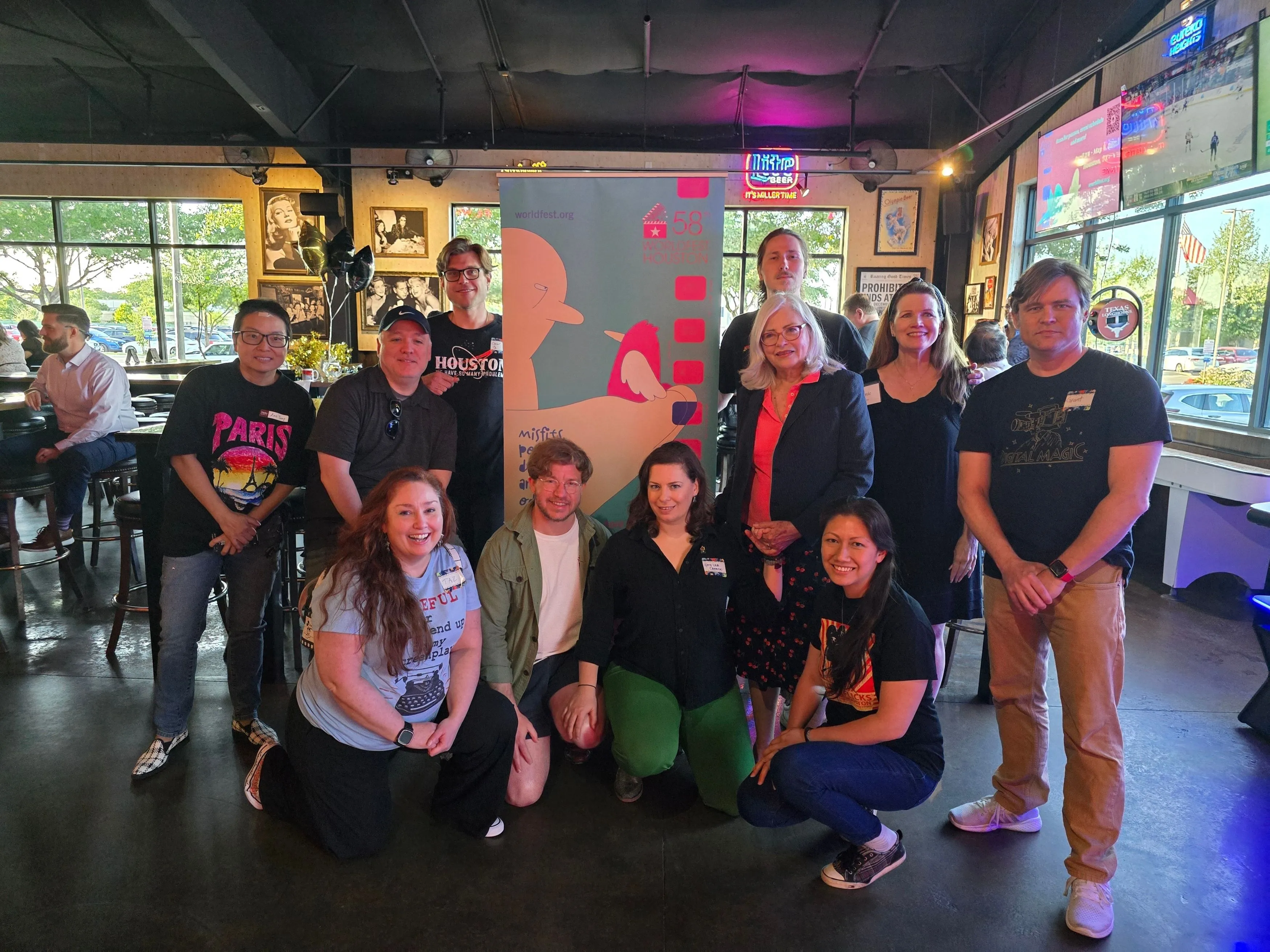 Group of diverse men and women posing indoors around a colorful WorldFest Houston banner, smiling at the camera.