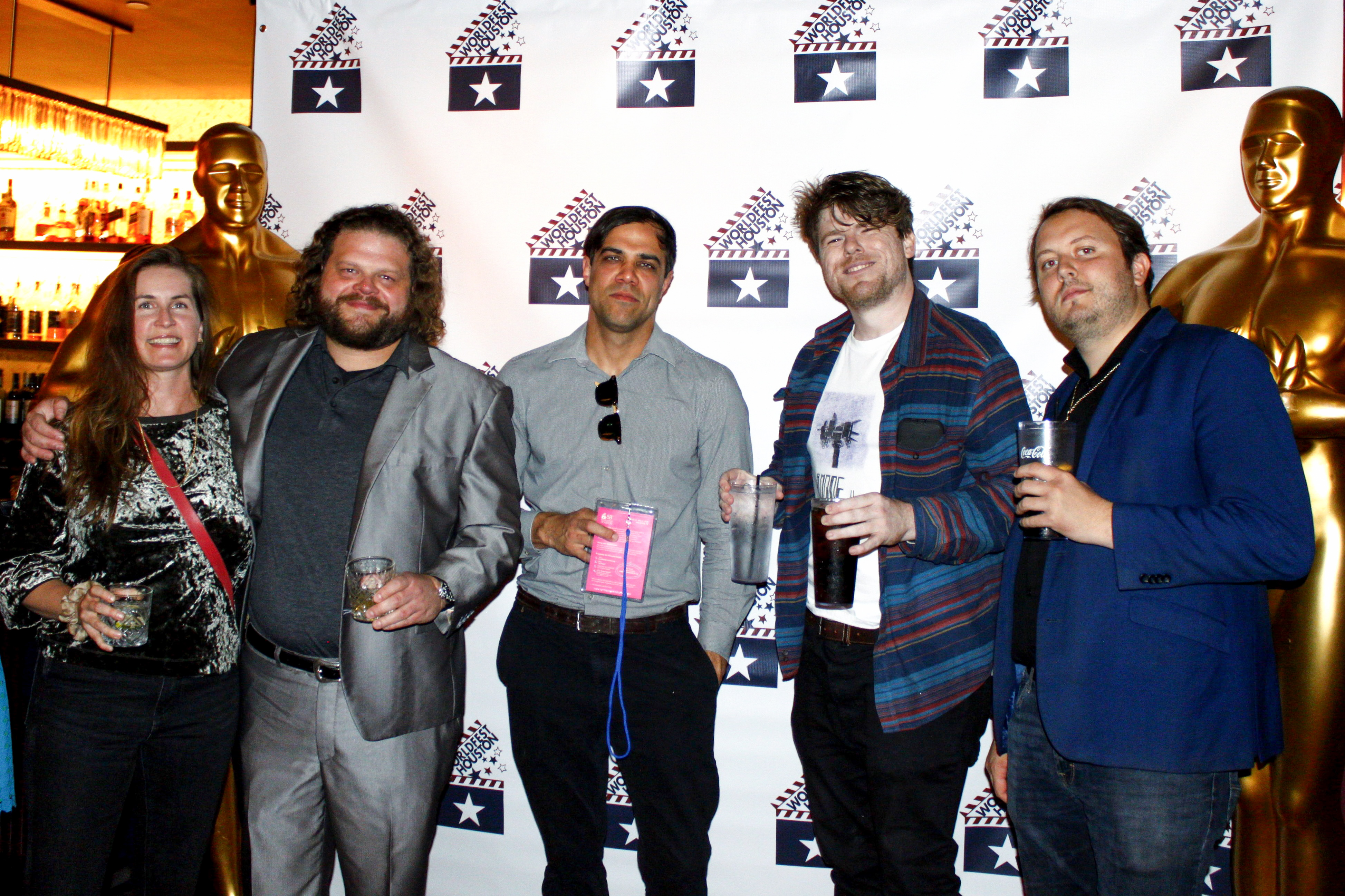 Five people posing with drinks in front of a WorldFest Houston backdrop, flanked by two large golden Oscar-style statues.