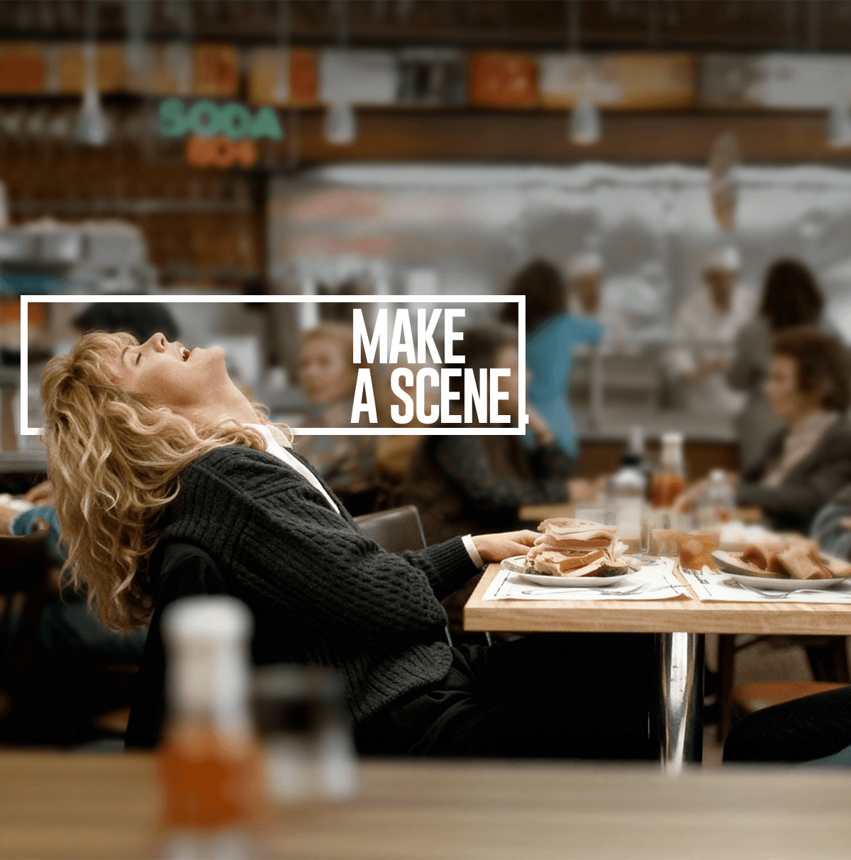 Woman with curly blonde hair leaning back and laughing at a diner table with sandwiches and condiments.