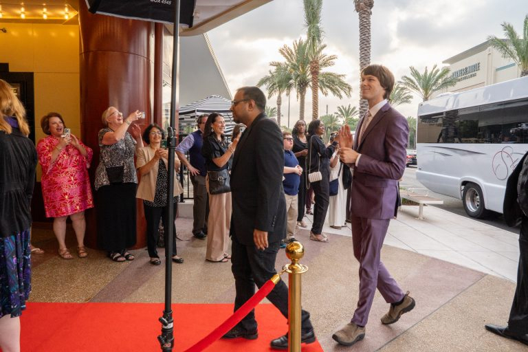 Two men walking on a red carpet outside a building with palm trees in the background; one man wears a black suit and the other a purple suit, while a group of people watch and take photos.