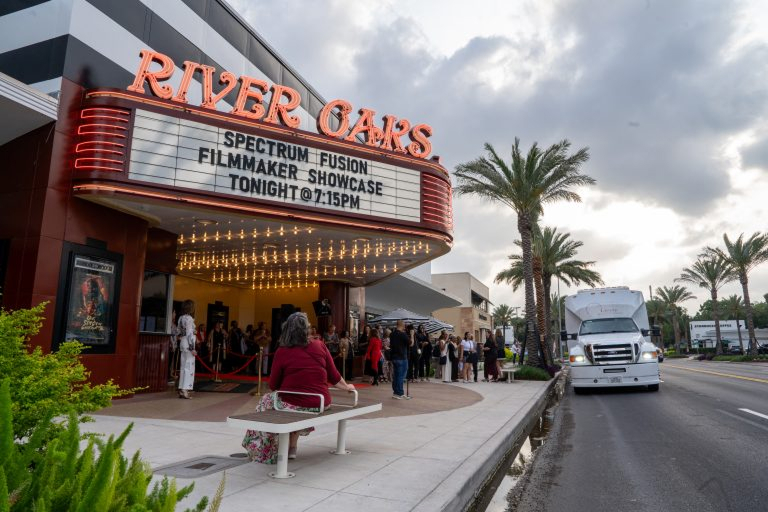 Exterior view of River Oaks theater with marquee announcing 'Spectrum Fusion Filmmaker Showcase Tonight @ 7:15PM', people gathered outside, woman in red sitting on a bench, and a truck parked on the street lined with palm trees under a cloudy sky.