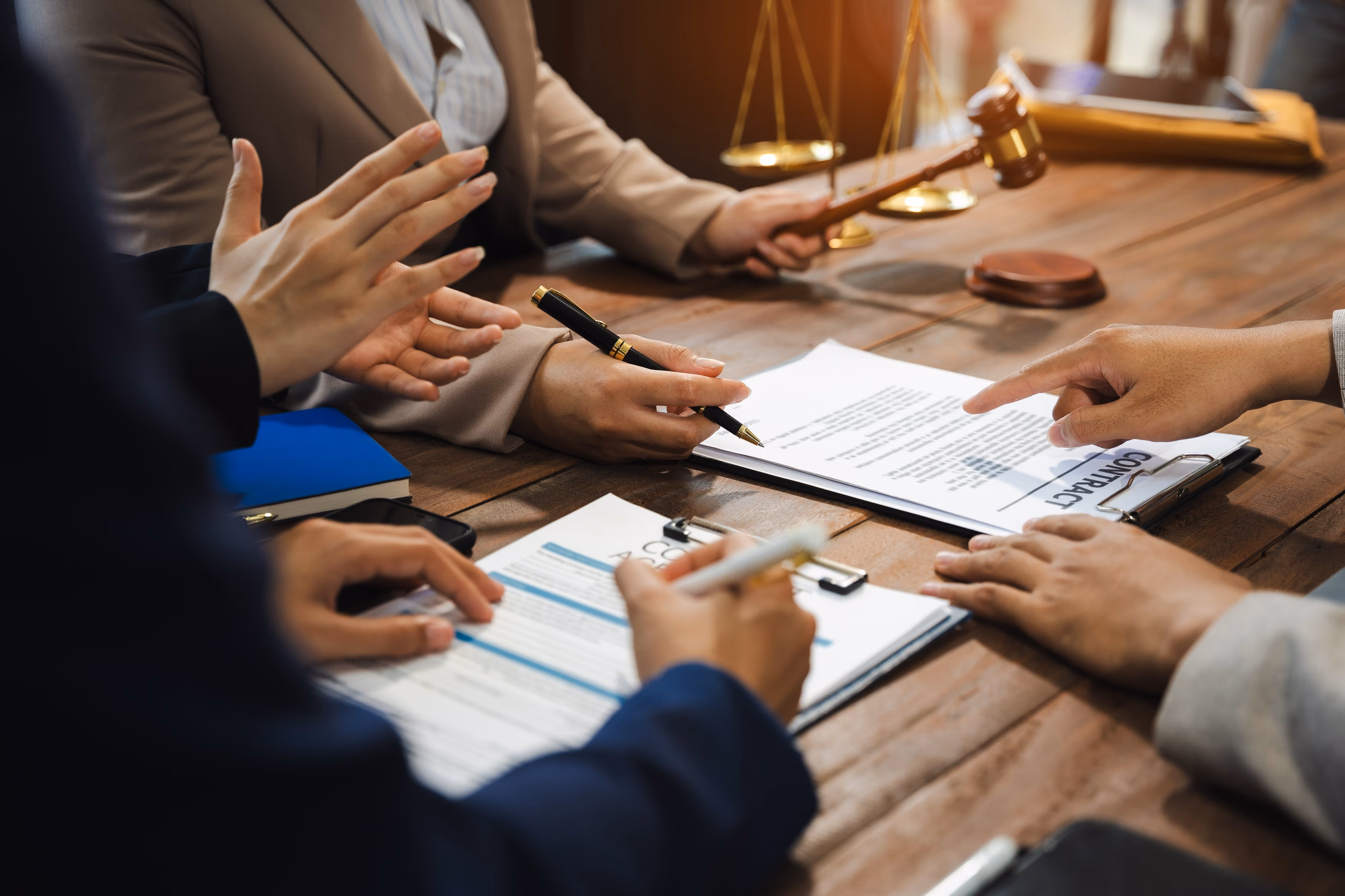 Several people in business attire are sitting at a wooden table discussing legal documents 