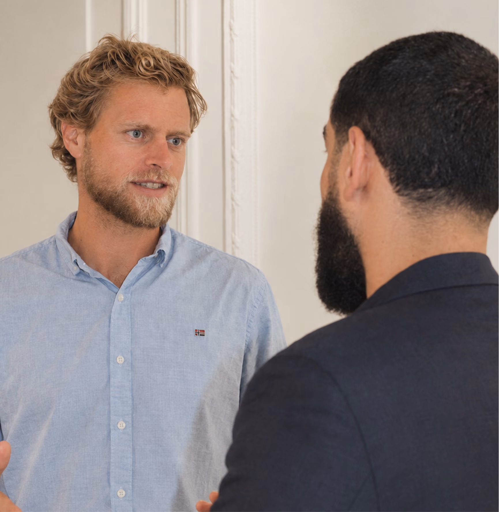 Un homme à chemise bleue clair parle avec un homme à barbe et veste sombre, dans un intérieur clair avec moulures murales.