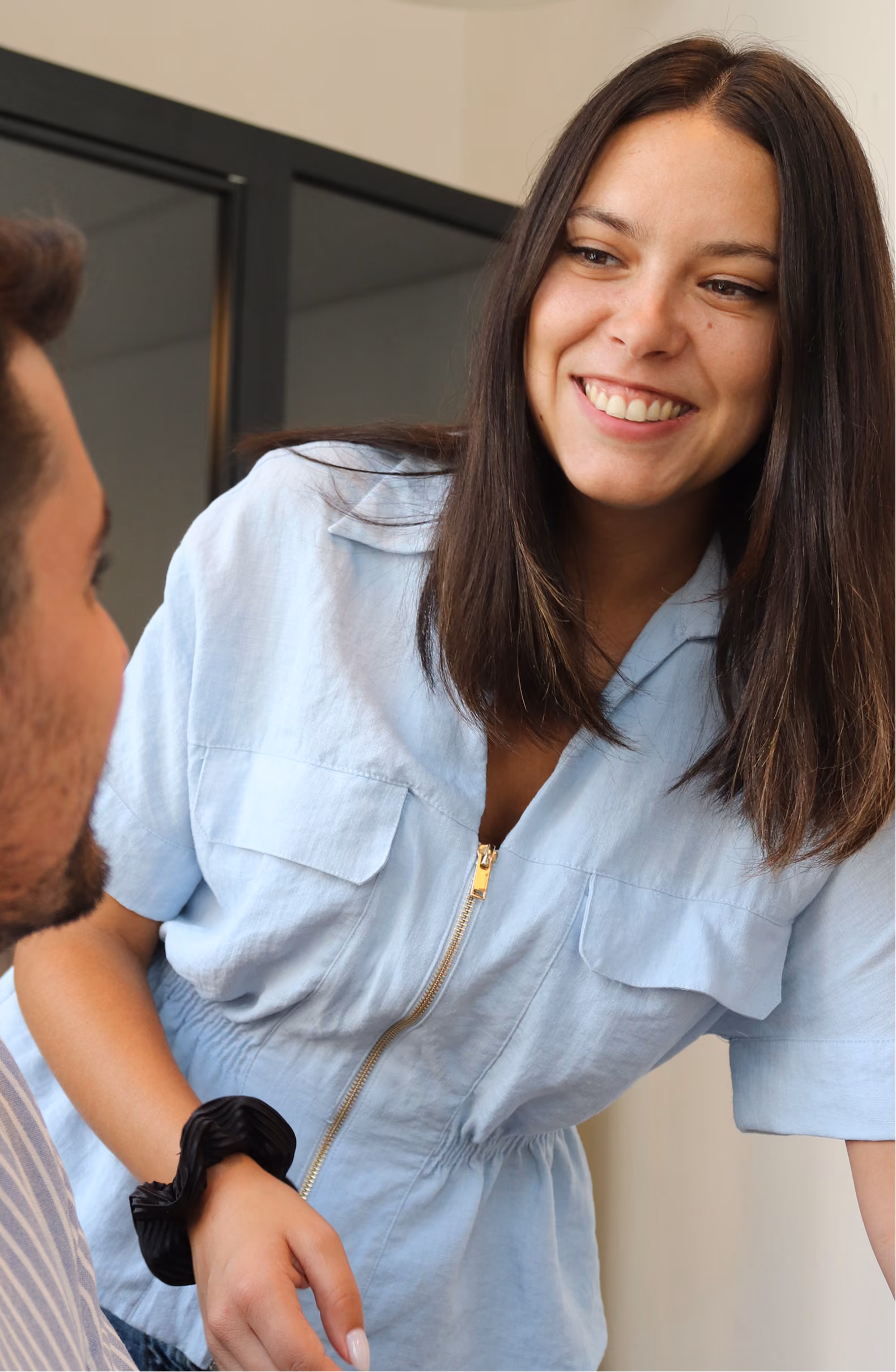 Jeune femme souriante en chemise bleue claire avec fermeture éclair, parlant à un homme assis en avant-plan dans un intérieur moderne.