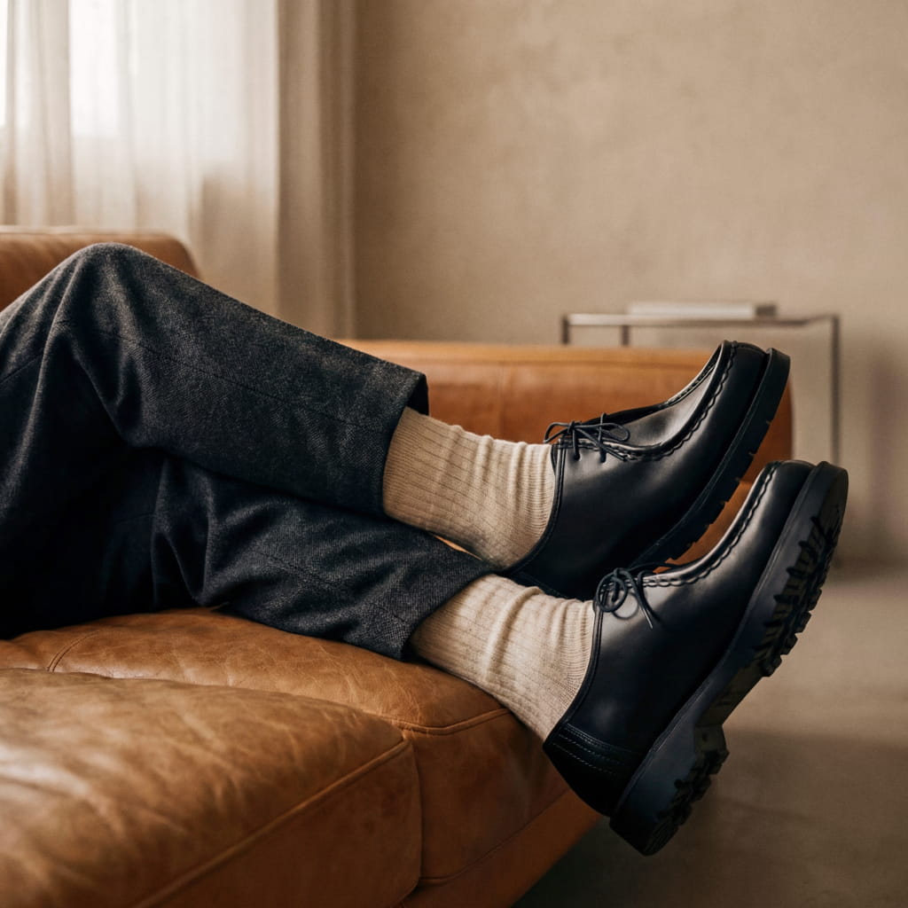Black leather lace-up shoes worn with beige socks and dark trousers, resting on a brown leather sofa in a minimal interior