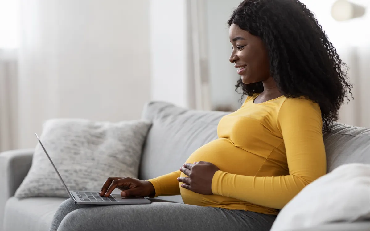woman sitting on her couch with her laptop, holding her pregnant belly, on  virtual lactation call with SimpliFed