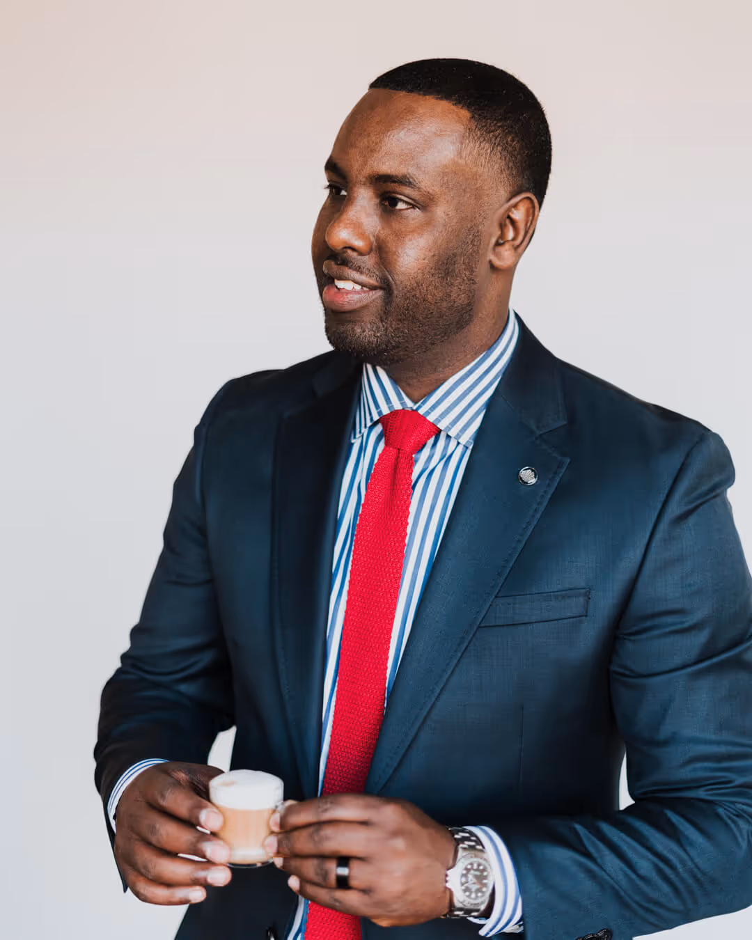 Image of Sacad Guled looking to the left and holding an espresso while wearing a navy suit and red tie with a pinstriped button-up shirt.