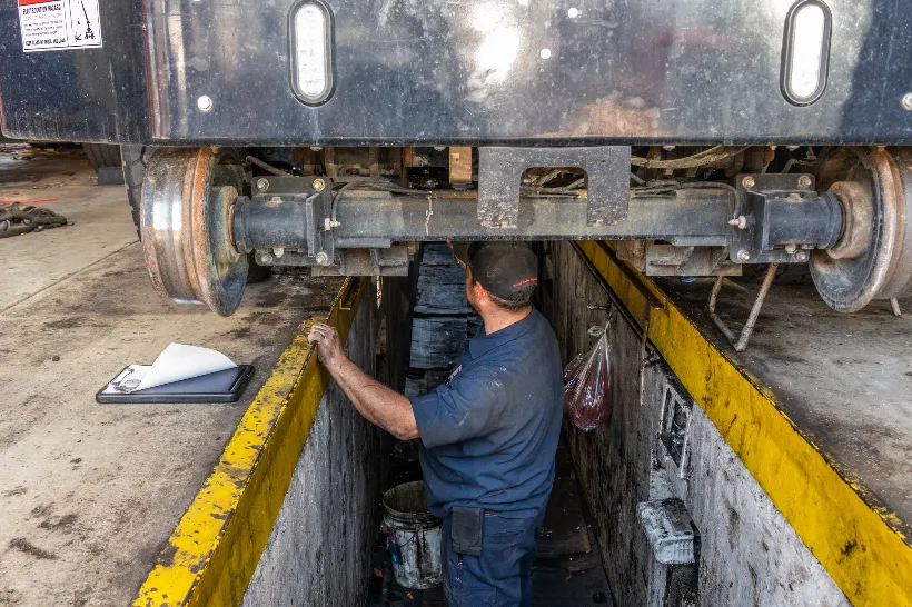 Technician performs fleet maintenance under a raised trailer, checking suspension and axle components in a service pit.