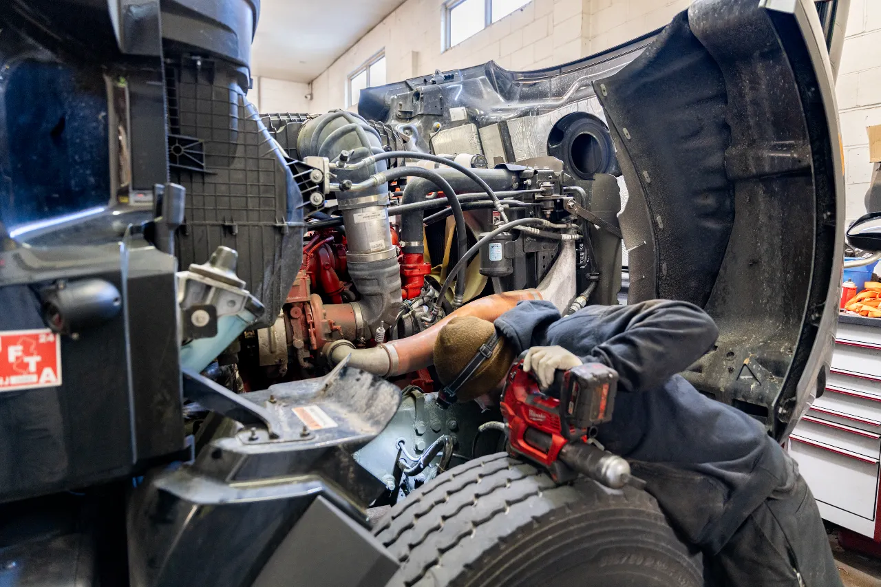 Mechanic performing preventive maintenance on a truck engine with a power tool and protective gloves.