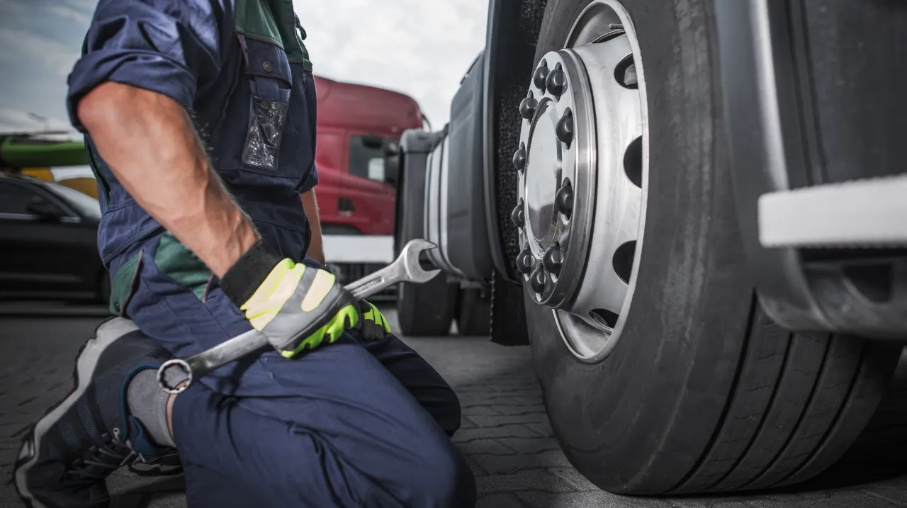 Technician performs preventive maintenance on a semi-truck wheel using a large wrench while kneeling.