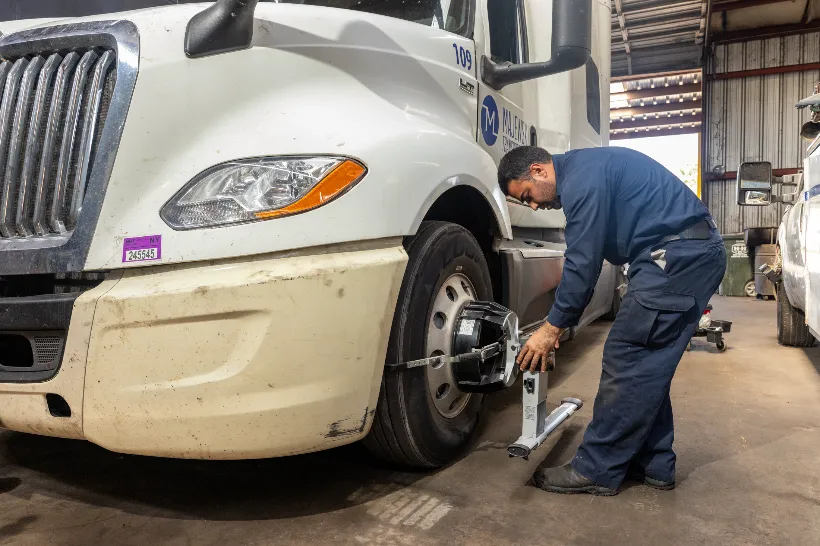 Technician performs fleet services by aligning a front wheel on a white semi-truck using precision equipment.