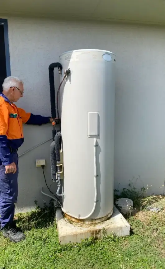 A technician inspecting a residential electric hot water system, checking for maintenance needs or signs of wear around the base and pipework.