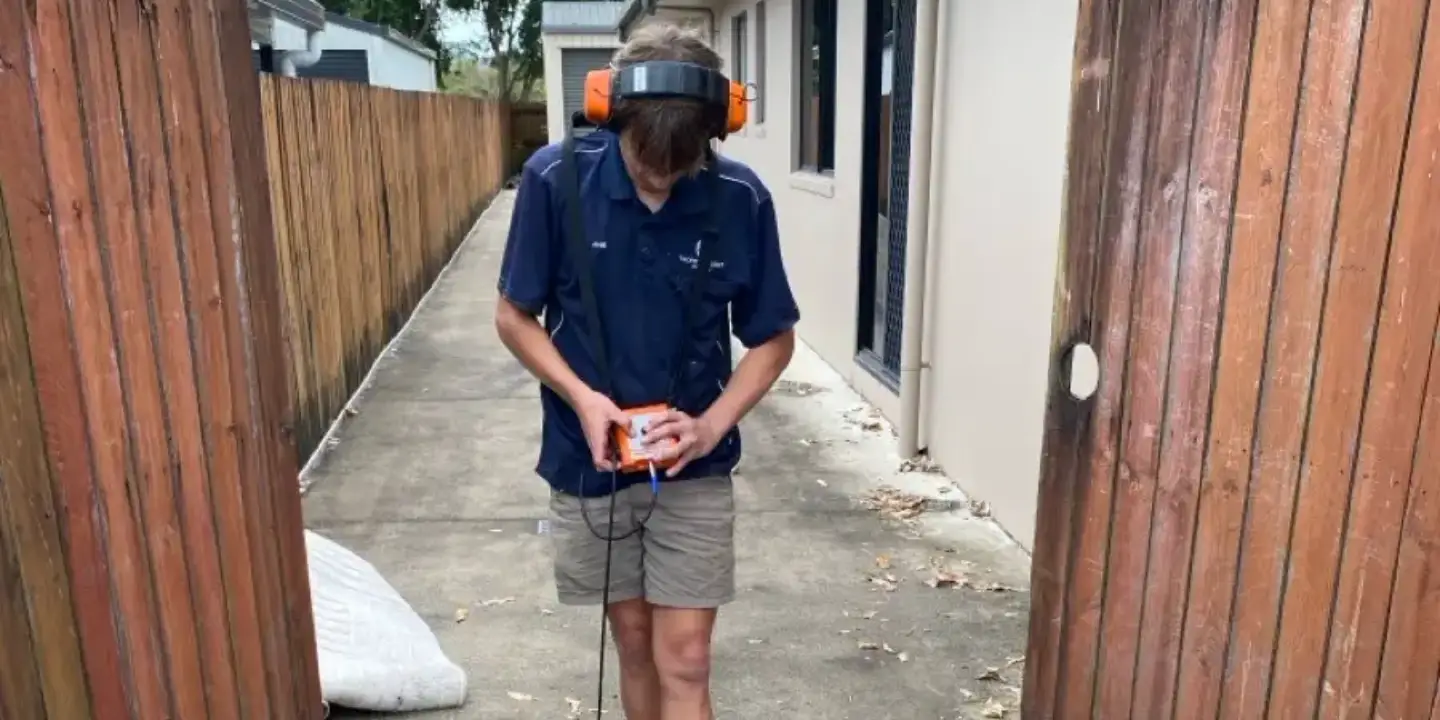 Tropical Coast Plumbing technician wearing safety earmuffs and visor operates leak detection equipment in a residential driveway.
