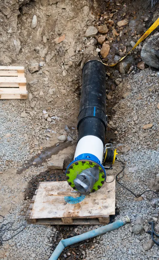 Large pipe with relining equipment installed in a dug-out trench on a construction site, showing the pipe opening secured with a green and blue flange.