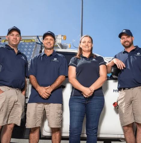 Members from Tropical Coast Plumbing stand smiling in front of a branded work vehicle parked outside their office.
