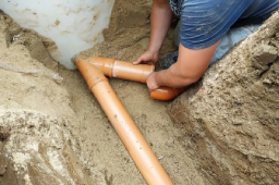 Plumber installing orange PVC sewer pipes underground, connecting fittings and ensuring proper alignment in a sandy trench.