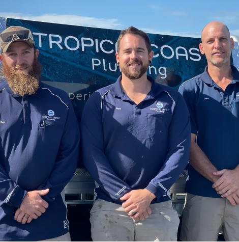 Members from Tropical Coast Plumbing stand smiling in front of a branded work vehicle parked outside their office.