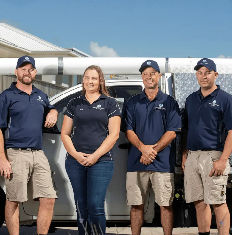 Members from Tropical Coast Plumbing stand smiling in front of a branded work vehicle parked outside their office.