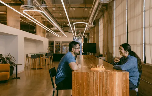 One of the many collaborative spaces found in the Discord offices — two employees are sitting across from each other, smiling and conversing over a chess game. They are seated at a long wooden table, various chairs and couches are behind them, as well as a refrigerator and snack area.