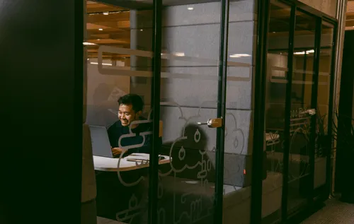 A Discord employee sitting in a private work booth, visible behind a glass door that is shut. He is working on his laptop and sitting on a comfy, fabric bench.