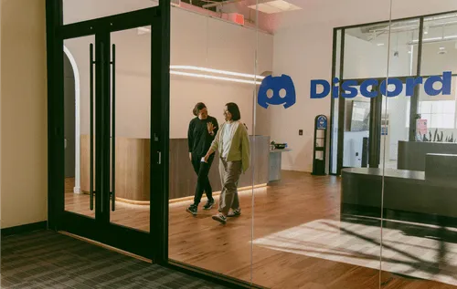 The entrance to the Discord office, with two employees pictured walking out and conversing. The Discord logo is visible from the front glass pane, along with a large glass door and reception area.