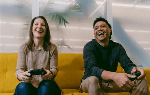 Two employees sitting atop a fabric couch, game controllers in their hands, both are visibly laughing as they play against each other in a video game. Behind them are various plants separated by a glass pane.