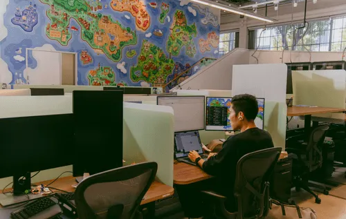 A Discord employee at work in the Discord office, sitting at his desk, surrounded by two monitors and a laptop. Behind it lays a mural containing popular references to various video games.