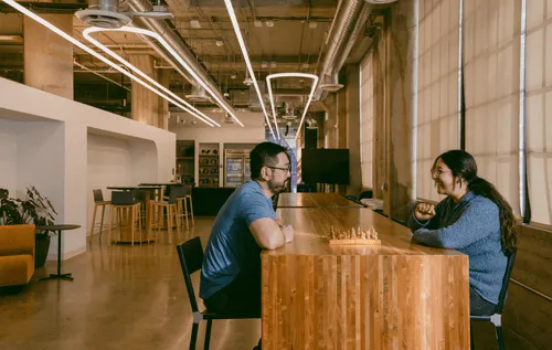One of the many collaborative spaces found in the Discord offices — two employees are sitting across from each other, smiling and conversing over a chess game. They are seated at a long wooden table, various chairs and couches are behind them, as well as a refrigerator and snack area.