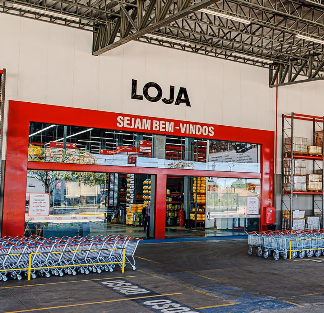 Entrance of a store with a large red frame and the words 'LOJA' and 'SEJAM BEM-VINDOS' above automatic glass sliding doors, with shopping carts lined up outside.