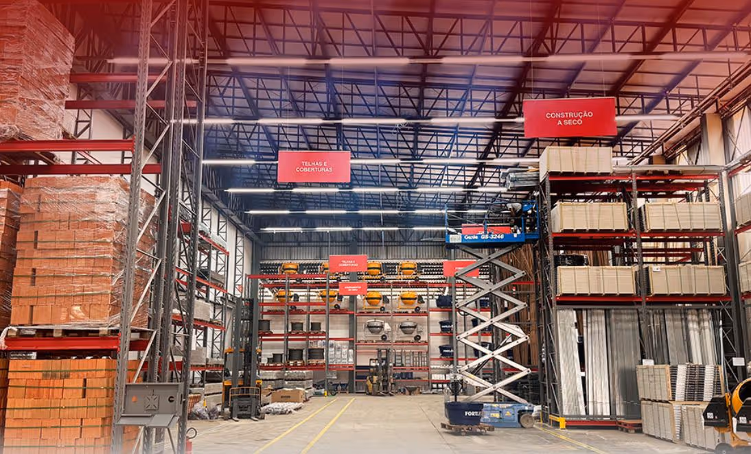 Interior of a warehouse with stacked construction materials including bricks, tiles, and equipment under high metal shelving with red signage.