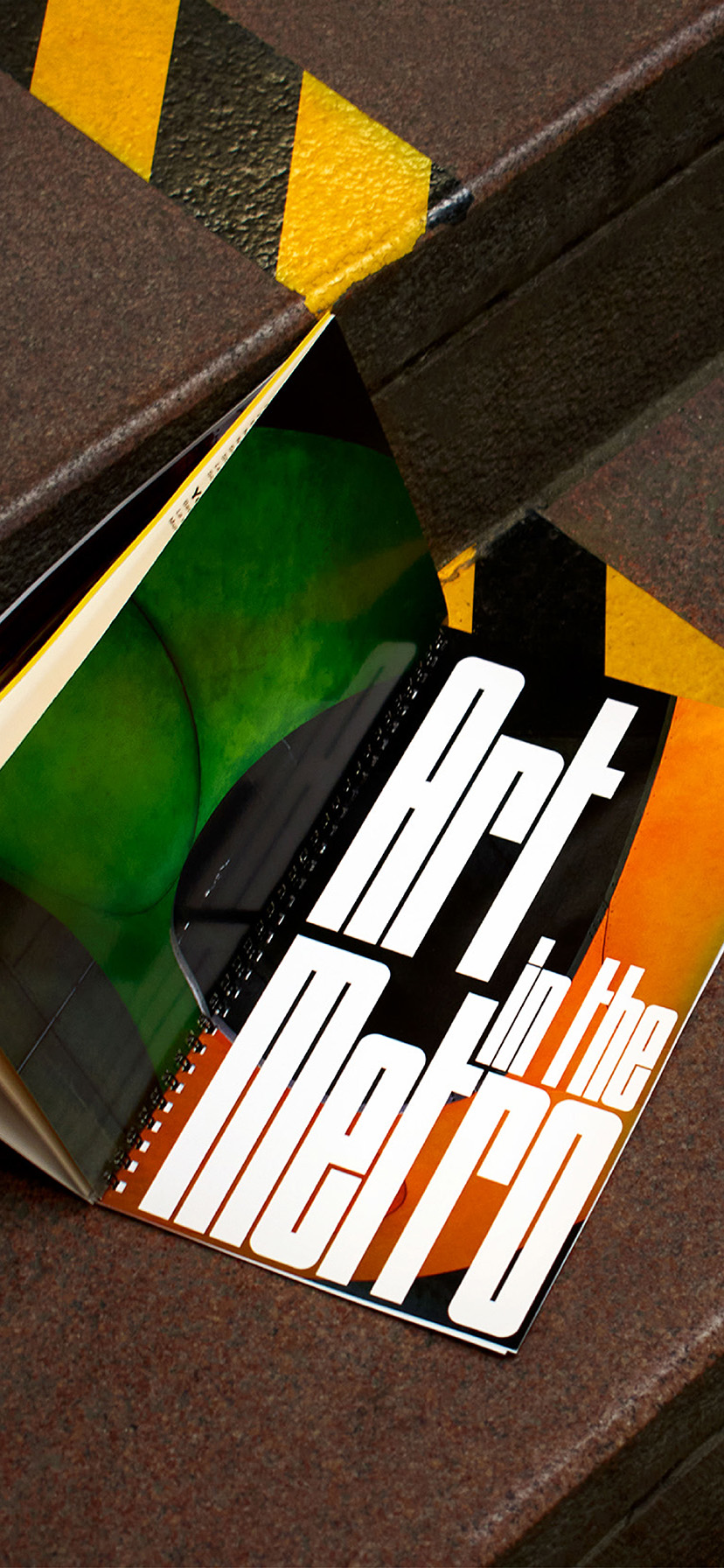 Book design opened and laid down flat on the stairs of a metro station (to show the back and front cover of the book). 3 steps can be seen, with the book placed on the middle step. A thick strip of black and yellow tape is on each step and the book is covering part of the tape on the second step.