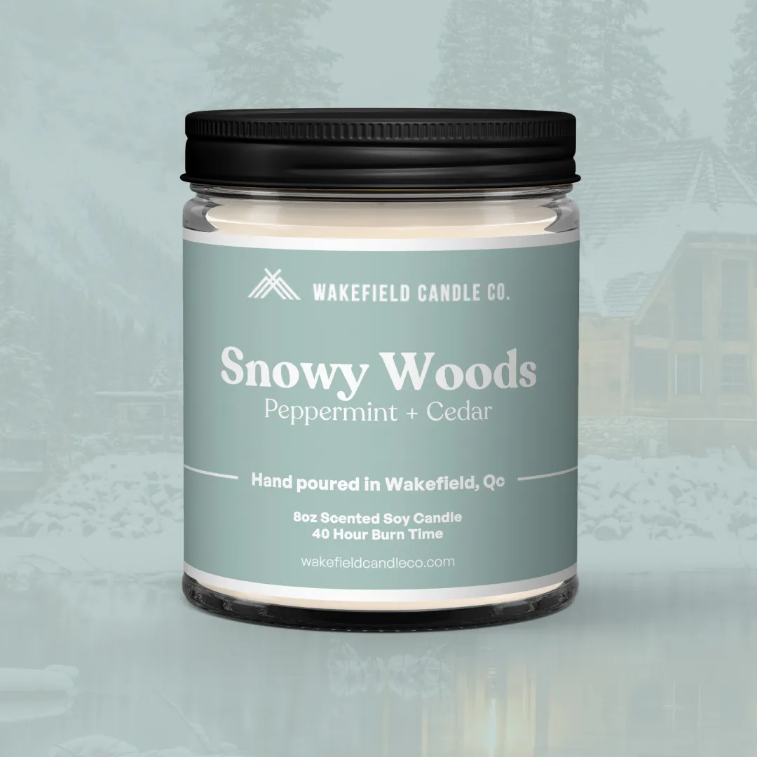 La bougie Forêt enneigée de Wakefield Candle Co. montrant l'avant de leurs bougies en traduction française. La bougie est posée sur un fond vert pâle discret où une image subtile apparaît, représentant un chalet douillet dans un paysage hivernal enneigé. 