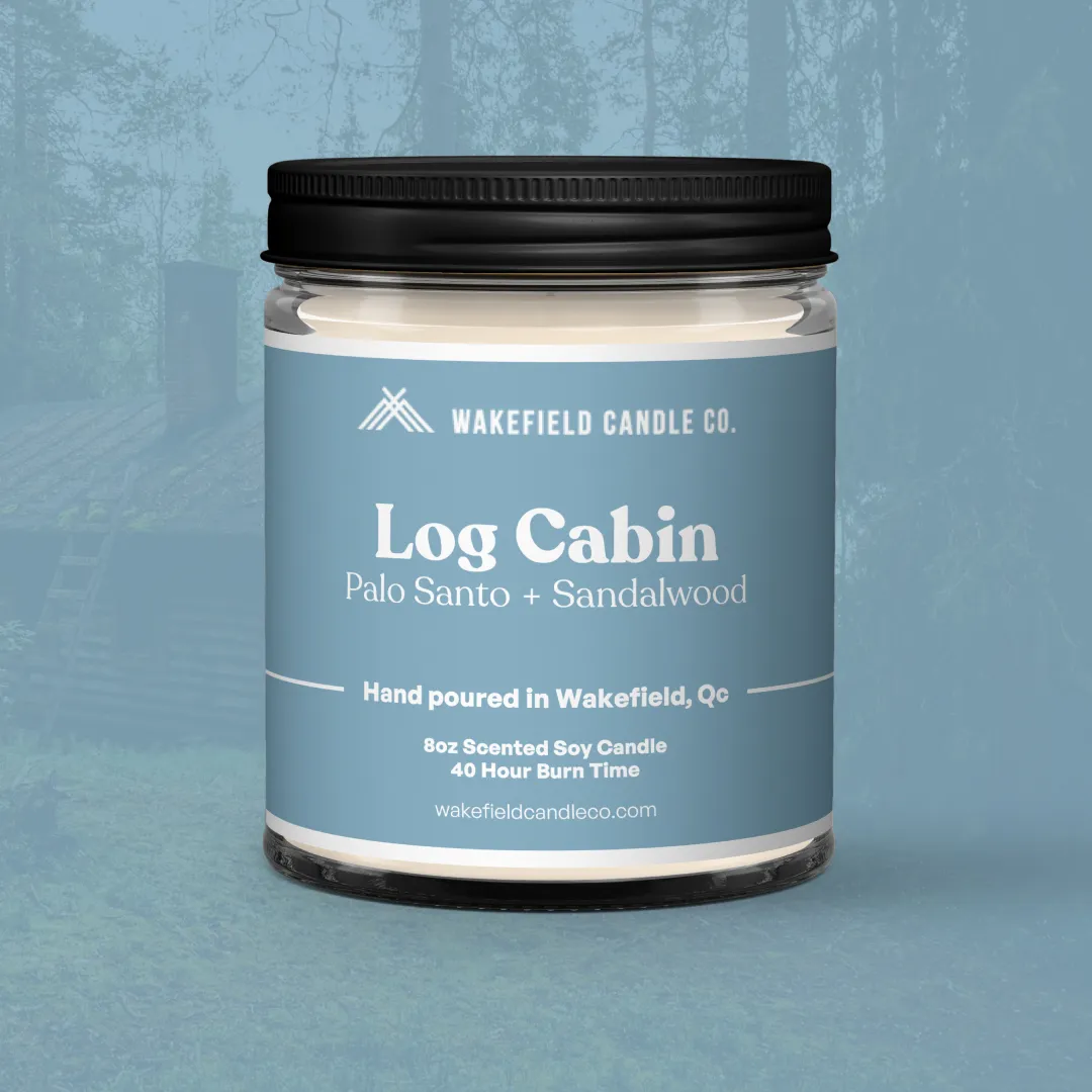 La chandelle Chalet en bois de Wakefield Candle Co. montrant le devant des étiquettes en traduction française. C'est une chandelle bleue posée sur un fond teinté de bleu qui présente un léger calque d'un chalet en bois en arrière-plan.