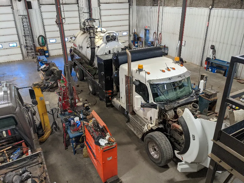 Engine repair service on white vacuum truck with hood open in large shop bay marked “1”.