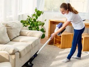 Woman vacuuming carpet near a beige sofa in a bright living room with a wooden side table and green plant.