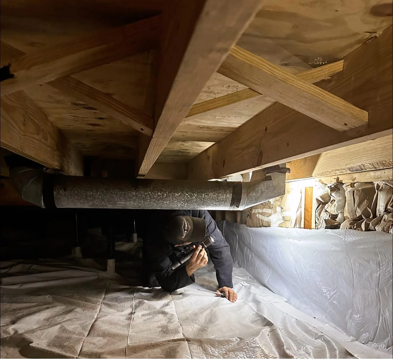 Person wearing a cap crawls in a crawl space under a wooden floor with a flashlight.