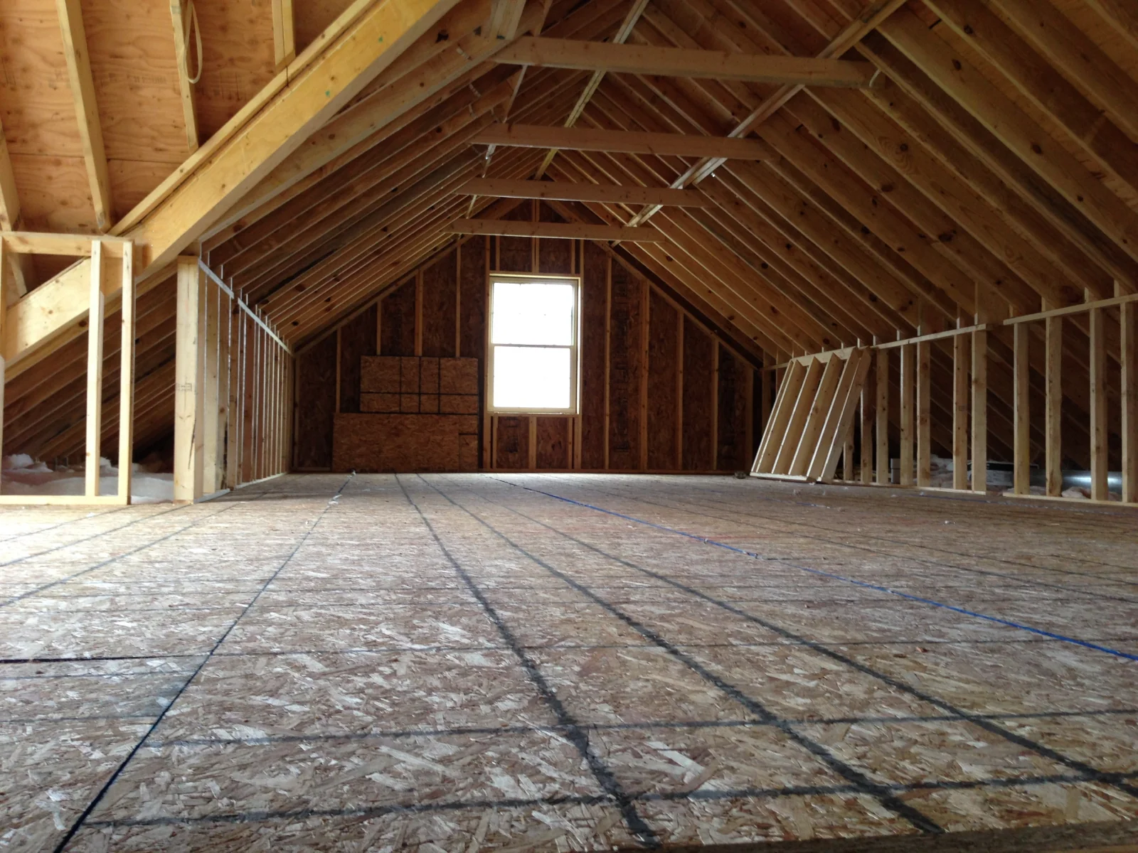 Empty attic space with exposed wooden beams, plywood flooring marked with grid lines, and a single window at the far end.