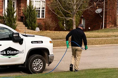 Technician wearing gloves spraying lawn treatment beside a white Black Diamond service truck in a residential neighborhood.