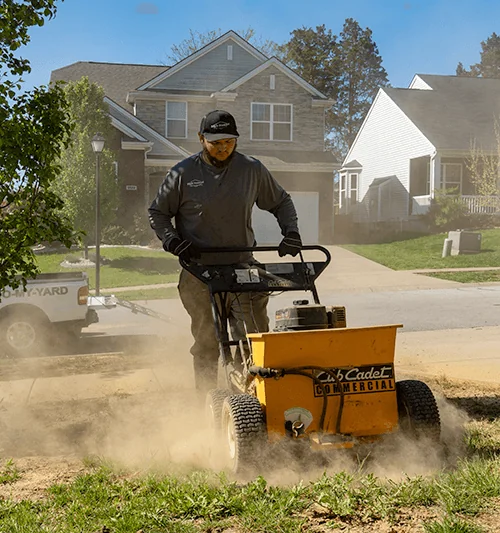 Man operating a yellow Cub Cadet Commercial lawn care machine on a dusty lawn in a suburban neighborhood.