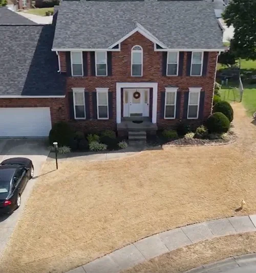 Two-story brick house with a gray roof, a dry brown lawn, and a black car parked in the driveway.