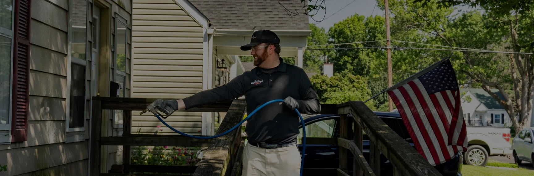 A pest control worker in black uniform and cap spraying insecticide near a house entrance with an American flag on the railing.