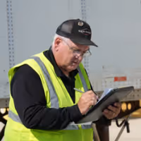 Man in a high-visibility vest and cap writing on a clipboard outdoors in an industrial area.
