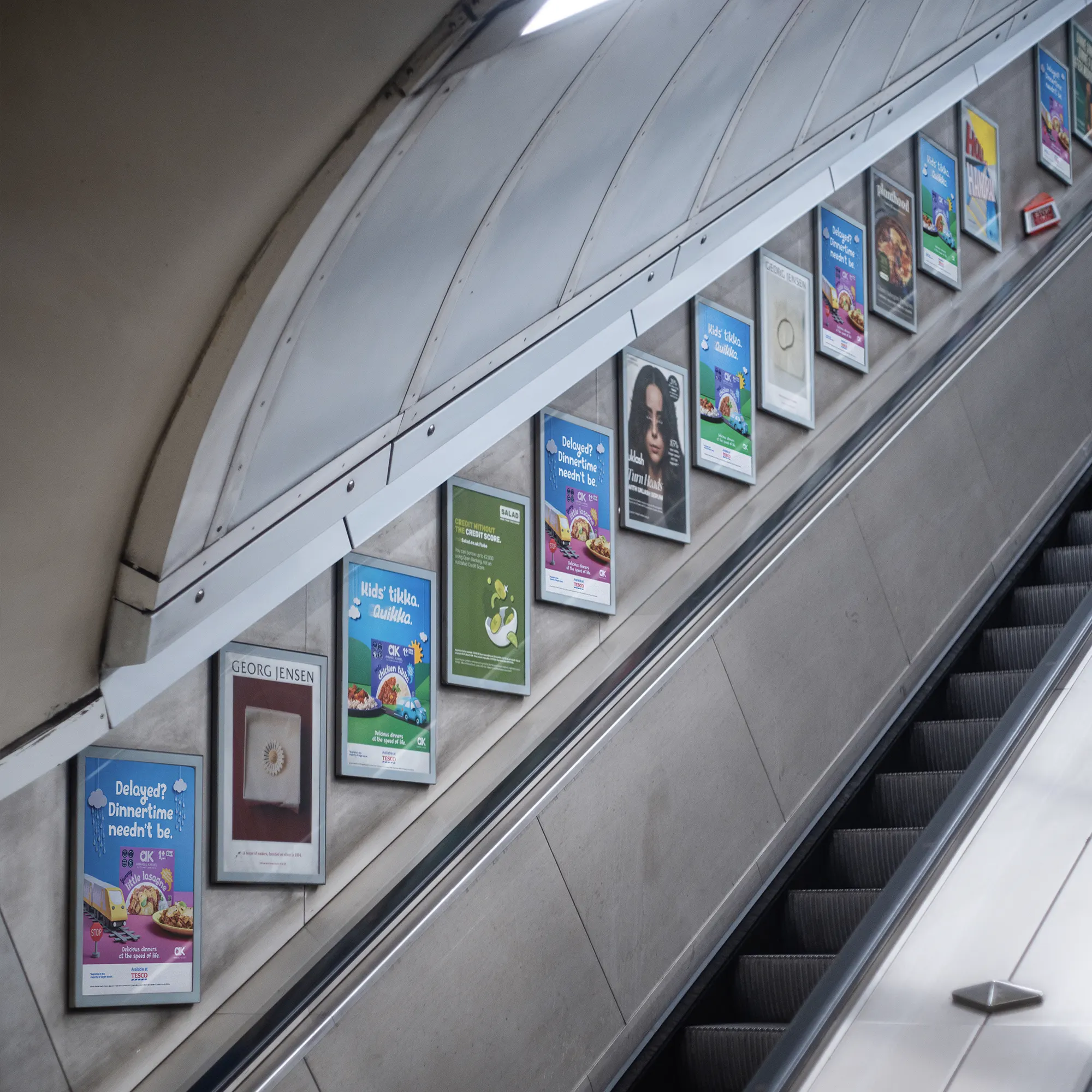 Escalator wall lined with framed advertisement posters for food, credit score, and jewelry brands.
