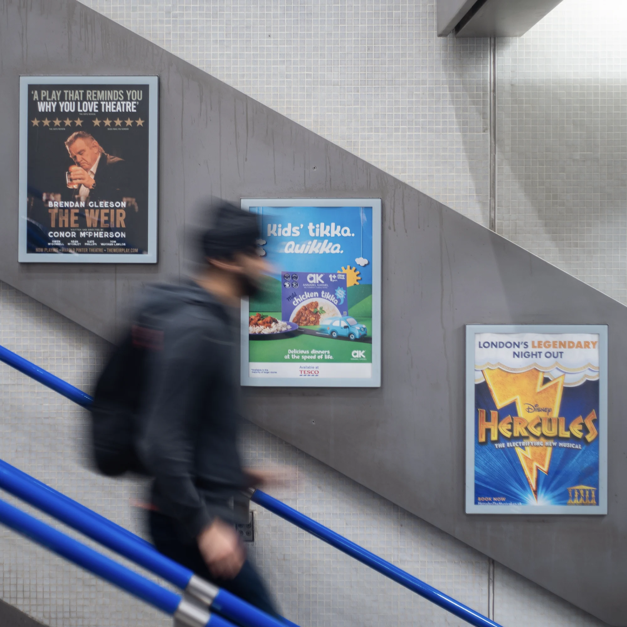 Blurred person walking up stairs with blue railings next to three framed posters on a tiled wall.