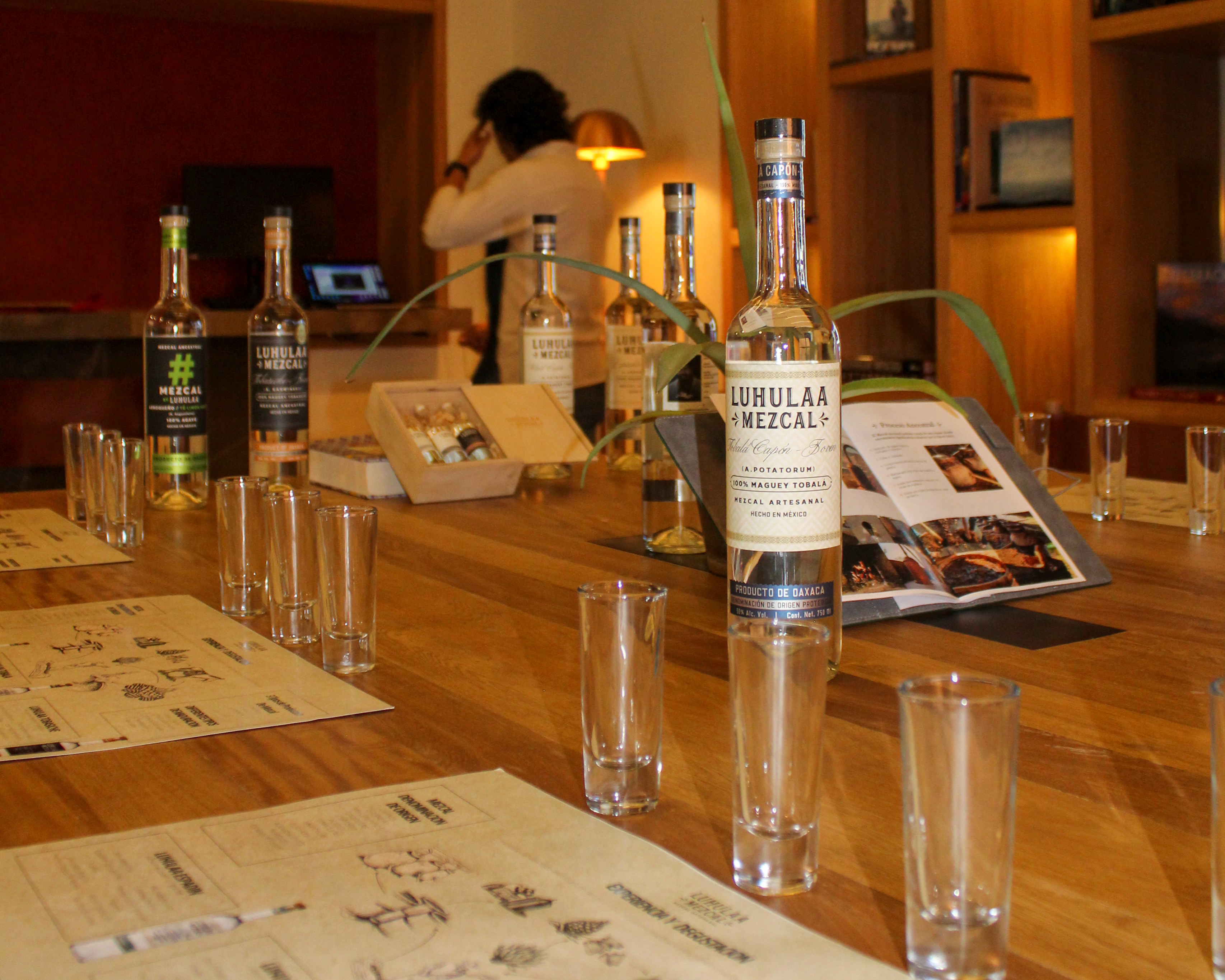 Mesa de degustación de mezcal con botellas de Luhlulaa Mezcal, vasos para shots y un libro abierto en un ambiente cálido y rústico.