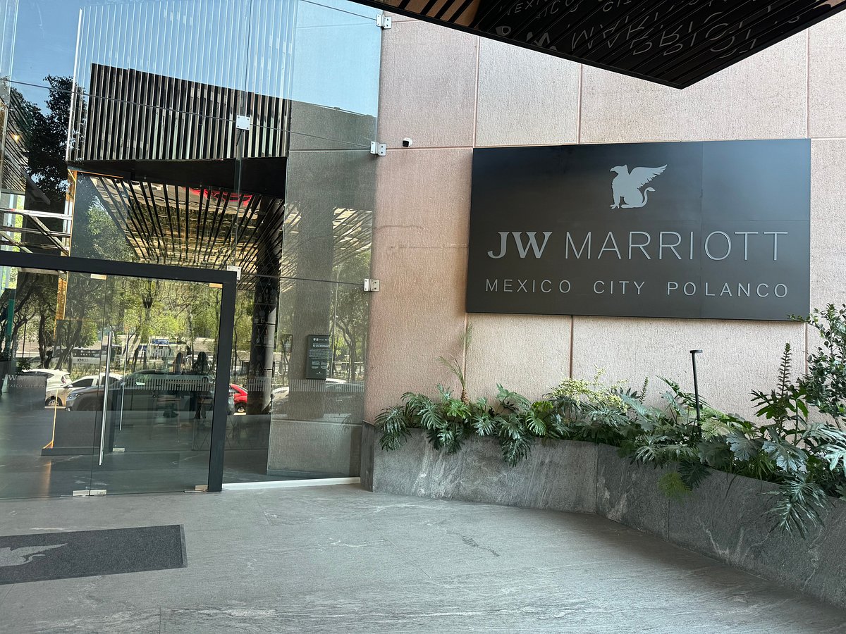 Entrada moderna del JW Marriott Mexico City Polanco con puerta de vidrio y un letrero negro en la pared junto a plantas decorativas.