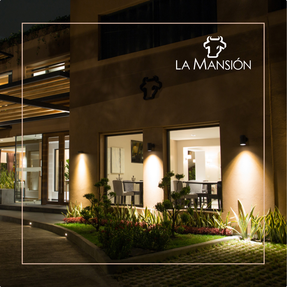 Fachada iluminada de restaurante La Mansión con jardín y ventanas que muestran mesas y sillas en el interior.