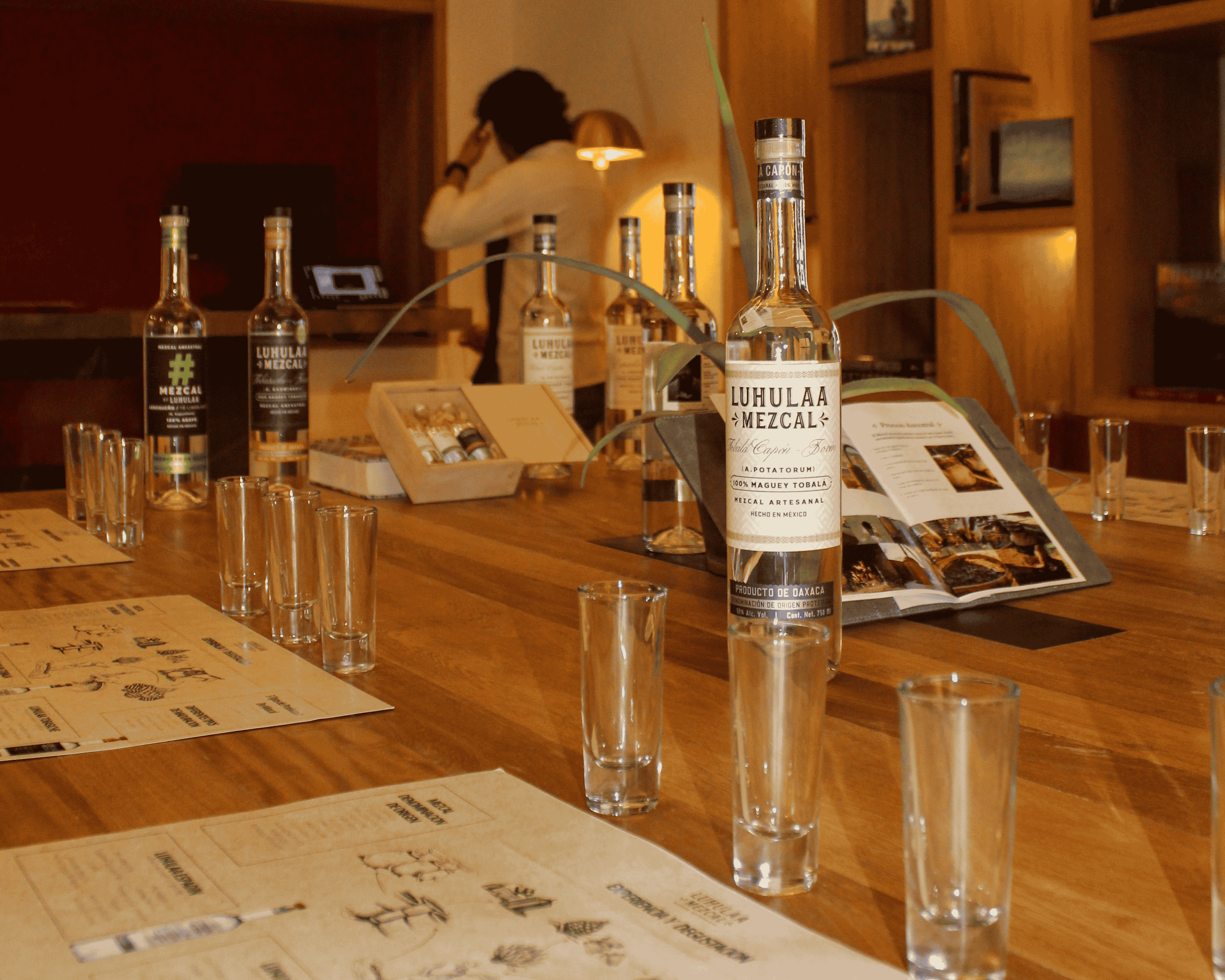 Botellas de mezcal Luhulaa y vasos alineados sobre una mesa de madera con menús y una persona desenfocada al fondo.